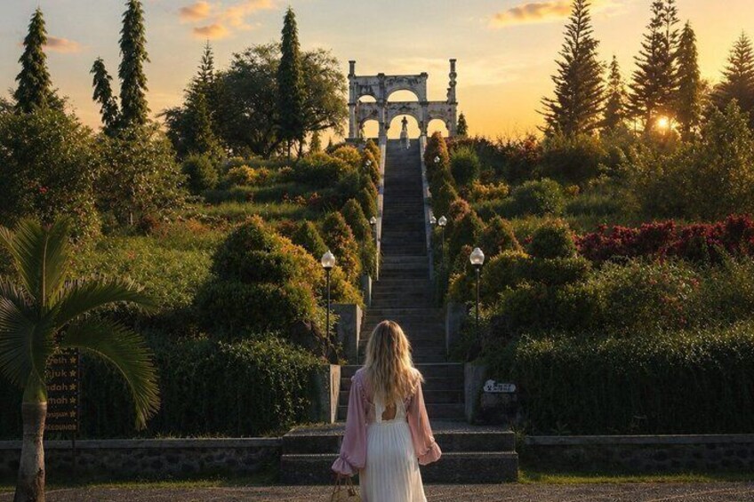 A woman in a white dress walking up a staircase towards a stone archway in a garden. 5
