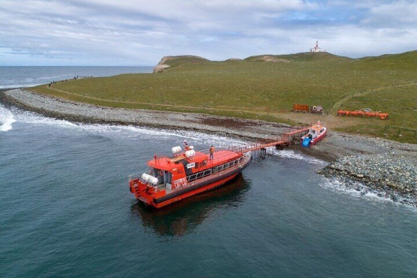 A red boat docked at a pier with a grassy hill and lighthouse in the background. 2
