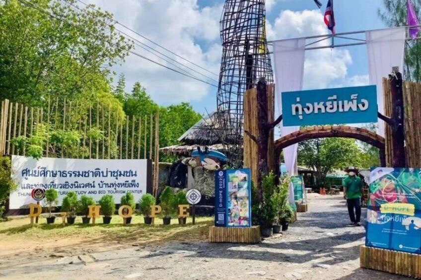 A rustic entrance with a sign in Thai, a bamboo fence, and a thatched-roof structure. 5