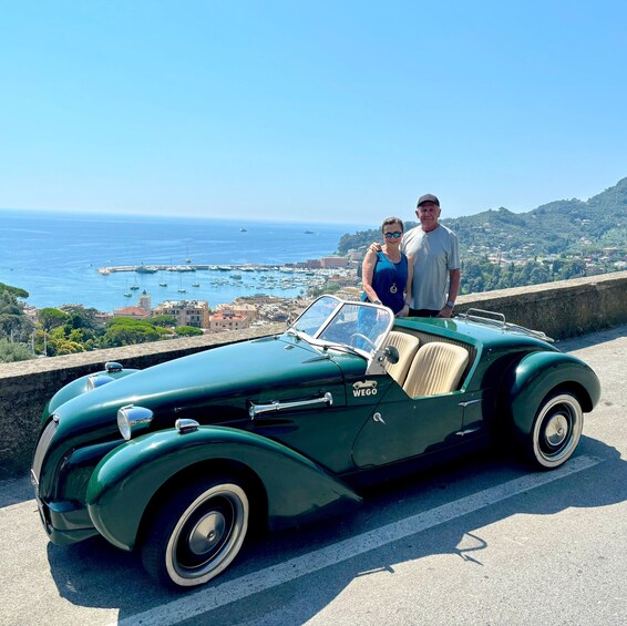 A vintage green convertible car parked on a coastal road with a scenic view of the sea and a town in the background. 2