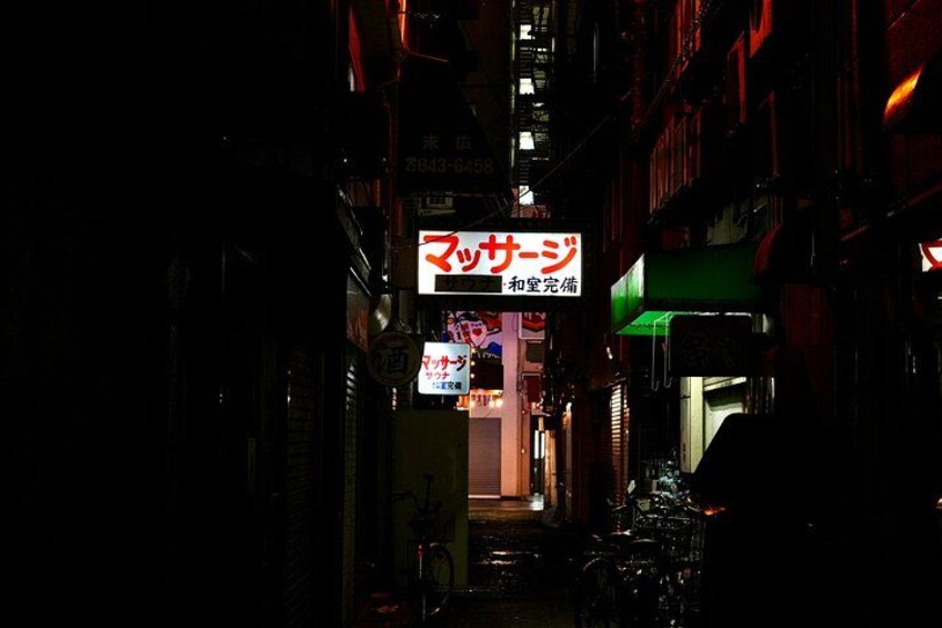 A narrow alleyway with illuminated signs, bicycles, and buildings. 1