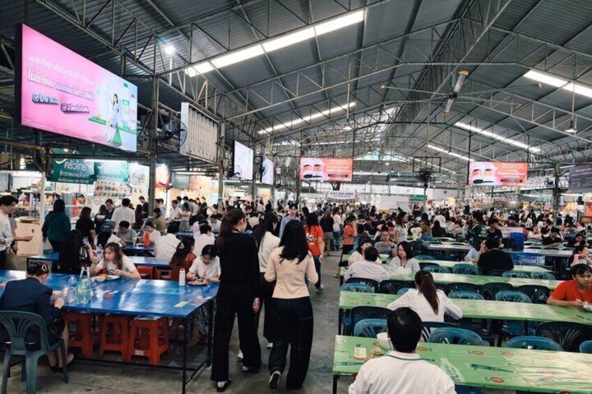 A crowded indoor event with people seated at tables, large digital screens, and a metal roof structure. 5