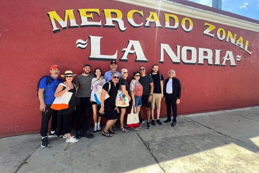 A group of people standing in front of a wall with the text 'Mercado Zonal La Noria'. 5
