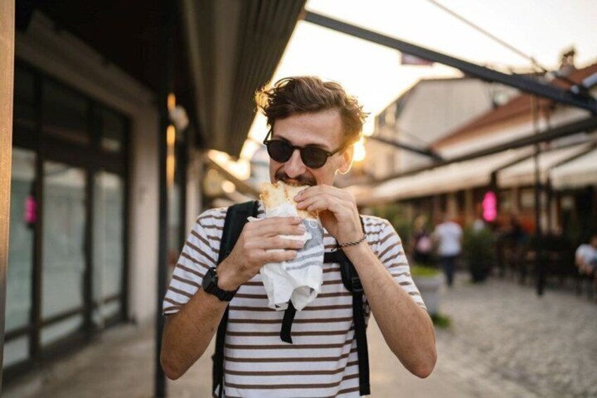 A person eating a sandwich outdoors, wearing a striped shirt and carrying a backpack. 1