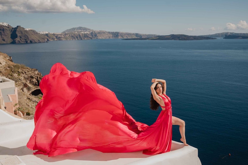 A person in a red dress sitting on a white structure by a body of water with mountains in the background. 2