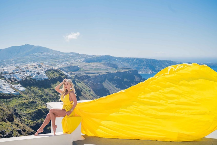 A person in a yellow dress sitting on a ledge with a yellow fabric draped beside them, overlooking a coastal landscape with buildings and hills. 5