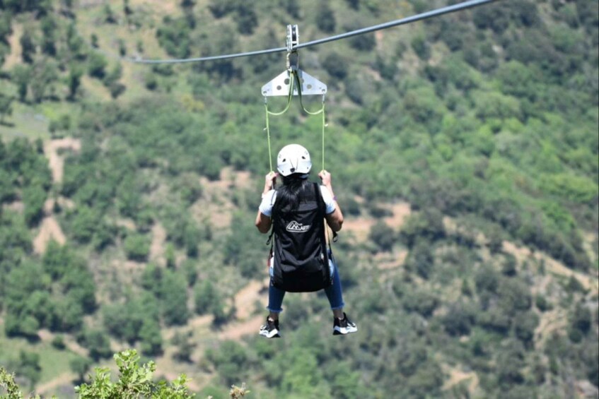A person suspended by a cable, wearing a helmet and harness, against a backdrop of a hilly landscape. 5