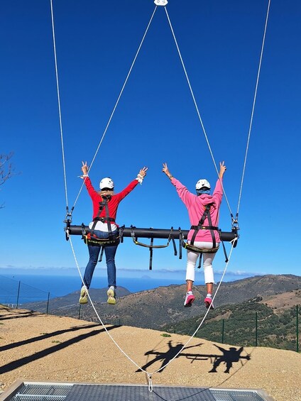 Two people suspended in the air by a harness system, with a clear blue sky and a mountainous landscape in the background. 4