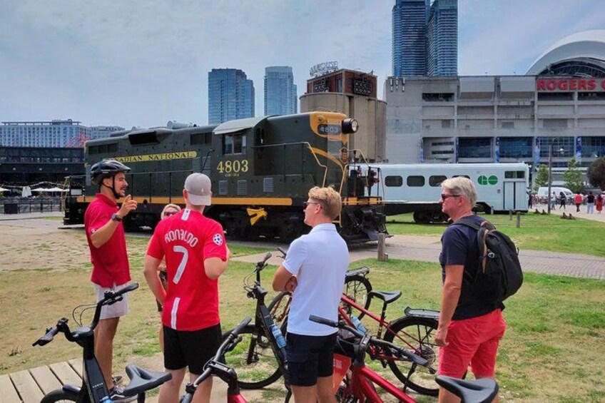 Een groep mensen staat bij fietsen en een trein, in een stedelijke omgeving met gebouwen op de achtergrond. 3