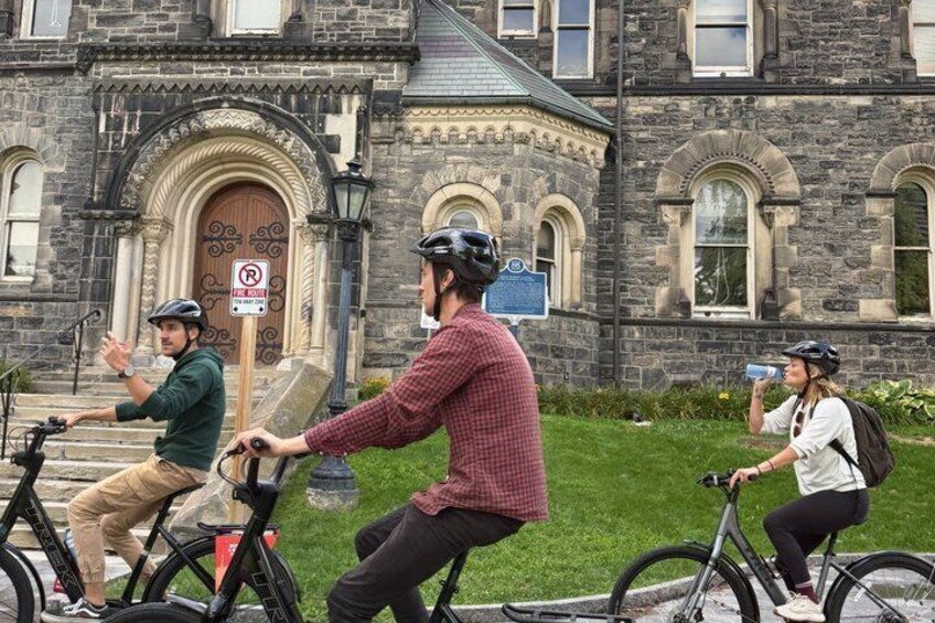 Three cyclists on a path in front of a historic stone building with a no parking sign. 4