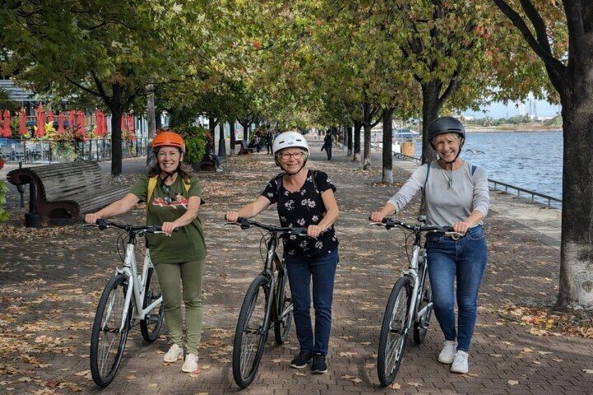 Three people on bicycles along a tree-lined path by a body of water. 1