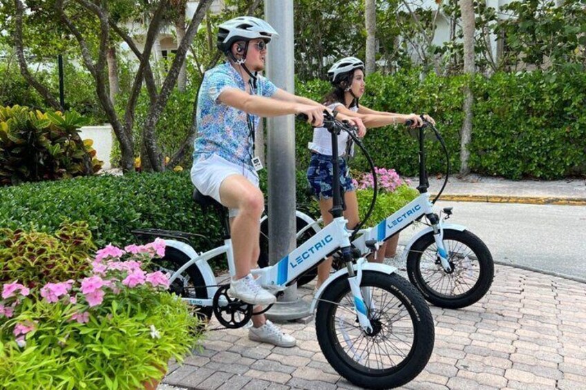 Two people riding electric bikes on a paved path with greenery and flowers. 1