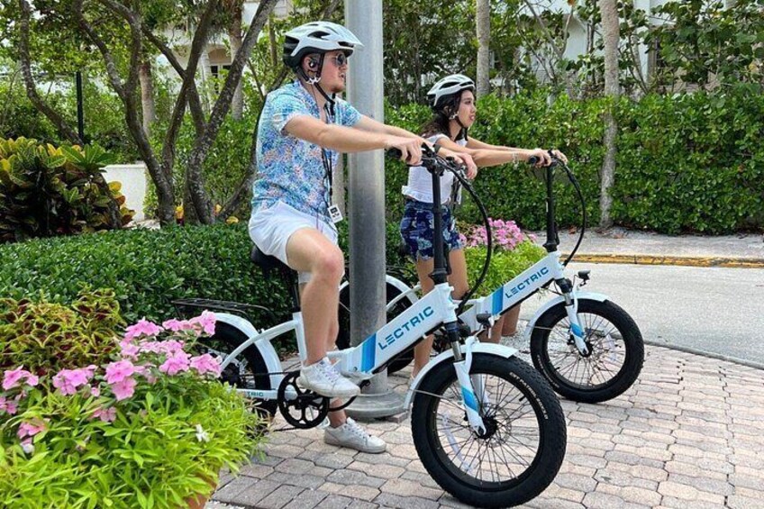 Two people riding electric bikes on a paved path with greenery and flowers around. 1