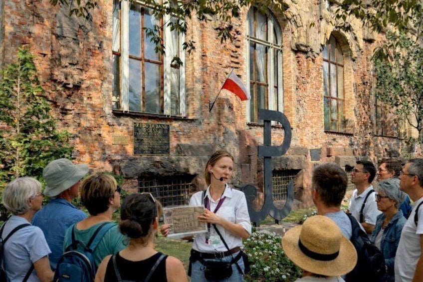 A group of people gathered in front of a historic building with a flag, a signboard, and a large letter 'P' structure. 3