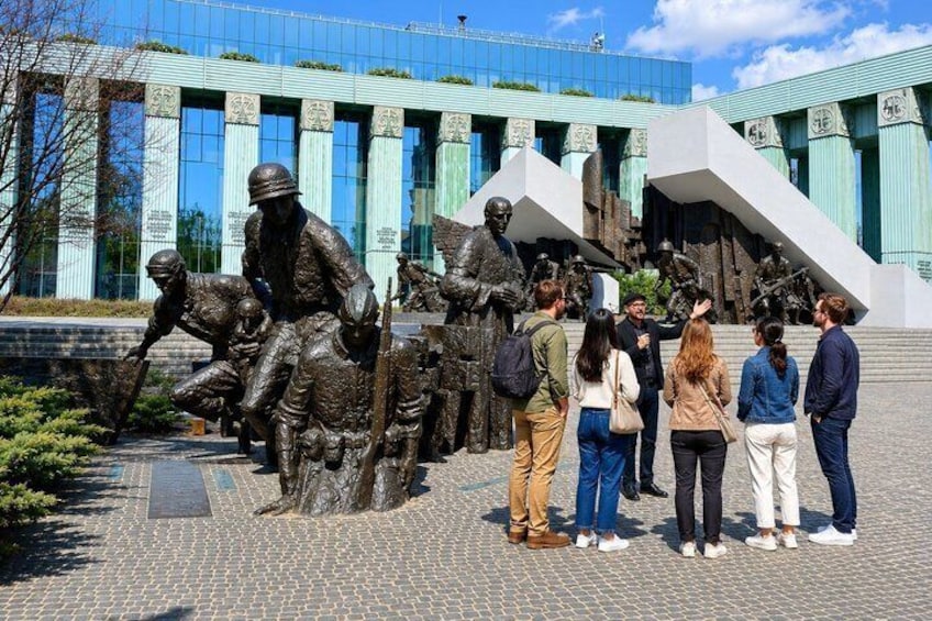 A group of people observing a bronze statue of soldiers in a public square with a modern building in the background. 1