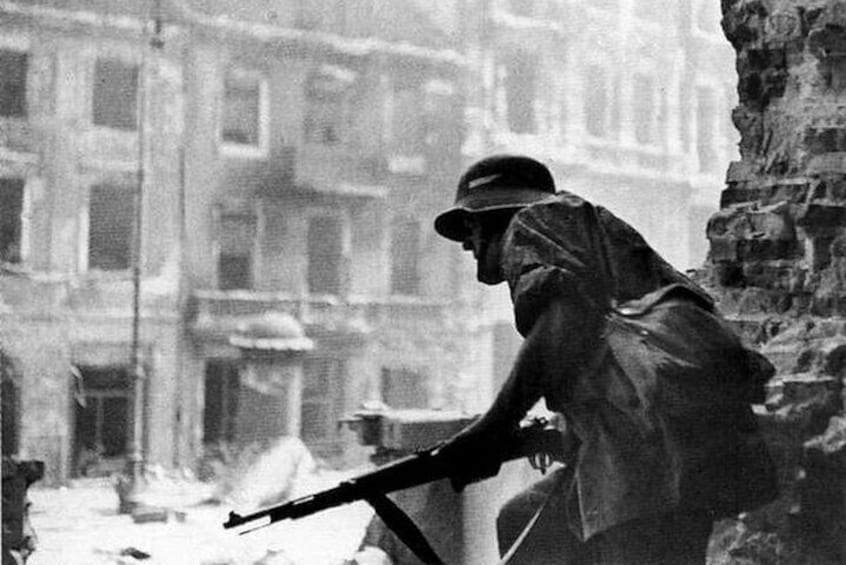 A person in a raincoat and hat holding a rifle, standing in a ruined urban environment with buildings in the background. 2