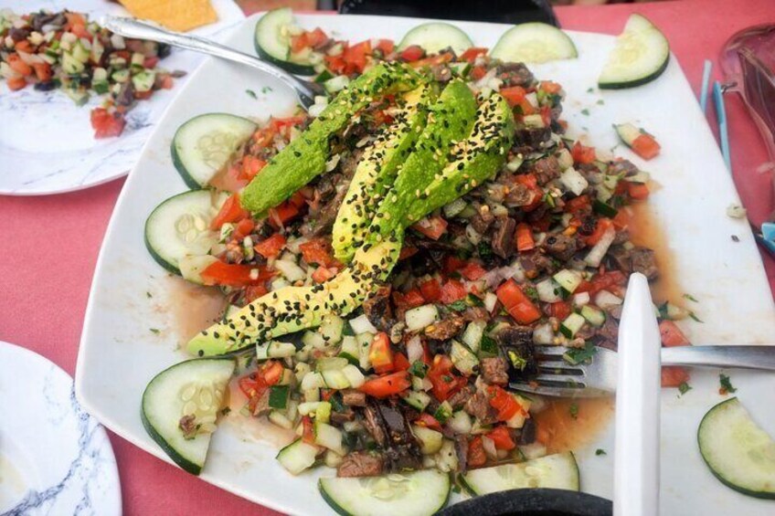 A plate of food with sliced cucumbers, diced tomatoes, and a piece of meat topped with a green vegetable and black seeds. 4