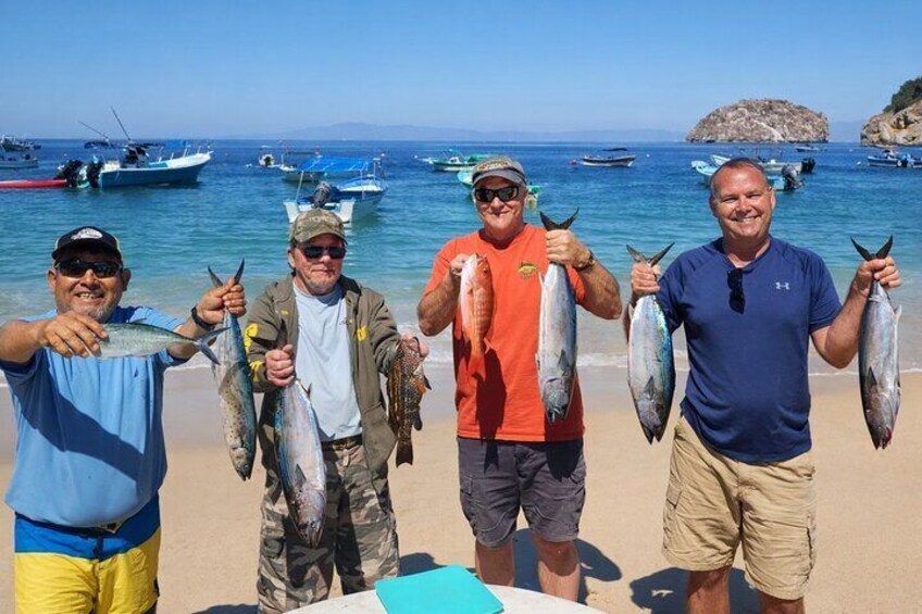 Five people on a beach holding fish, with boats and a rocky island in the background. 5