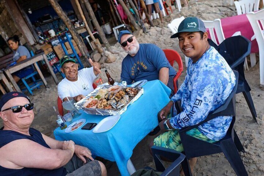 A group of people sitting at a table with food on a beach. 3