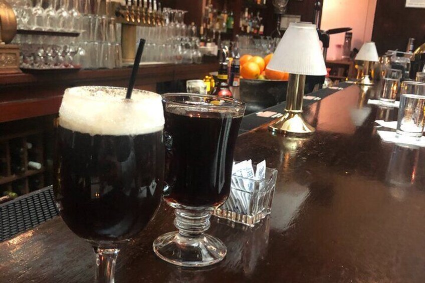 Two dark-colored drinks on a bar counter with a lamp and bottles in the background. 2