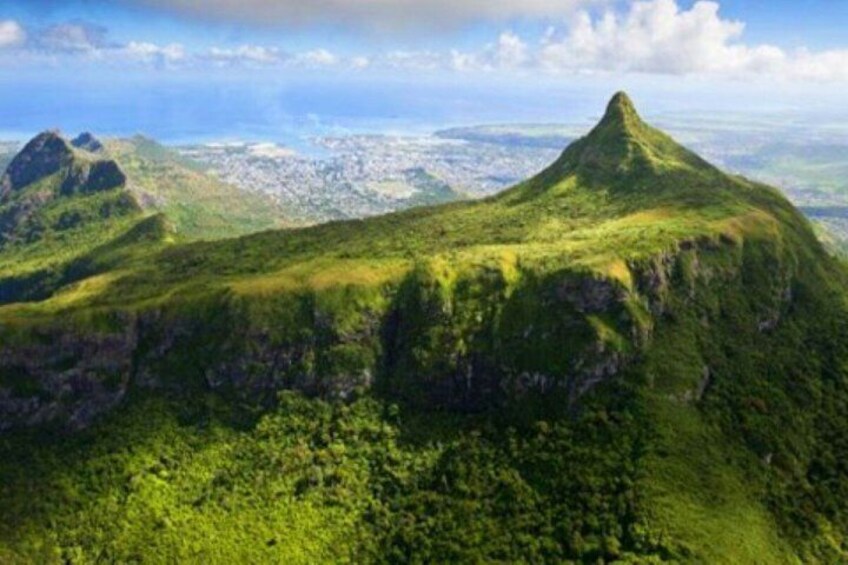 A lush green mountain with a distinct peak, surrounded by dense vegetation and a distant cityscape. 3