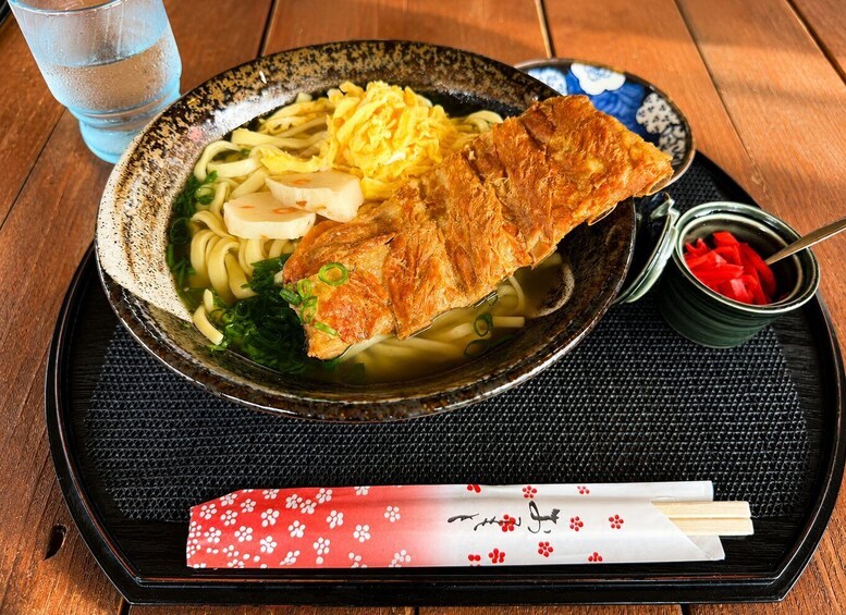 A bowl of ramen with a fried fish topping, sliced green onions, and a small bowl of red pickled vegetables on a wooden table. 5