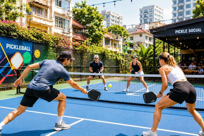 A pickleball game on an outdoor court with a colorful fence and buildings in the background. 1