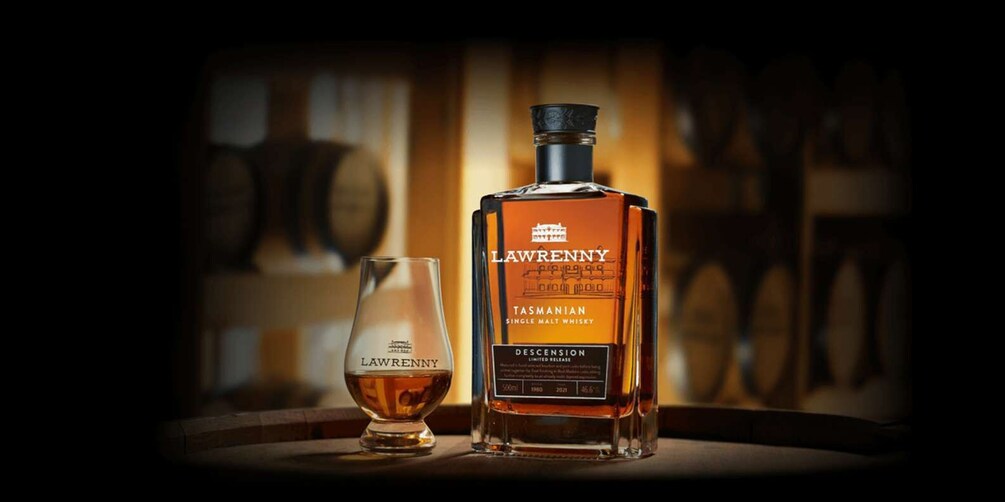 A bottle of Lawrenny Tasmanian Single Malt Whisky and a glass on a wooden surface with barrels in the background. 2
