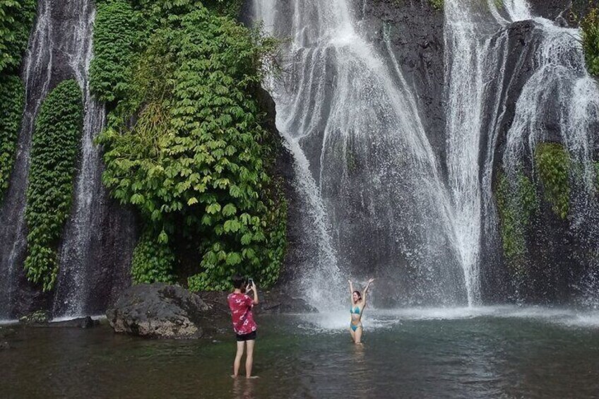 A waterfall with lush greenery and two people standing in the water. 5