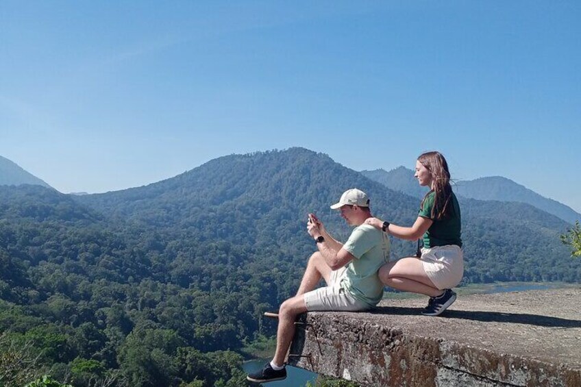 Two people sitting on a ledge overlooking a forested valley with mountains in the background. 3