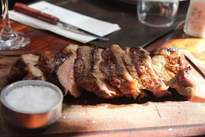 Sliced grilled meat on a wooden board with a salt container and a knife on a table. 5