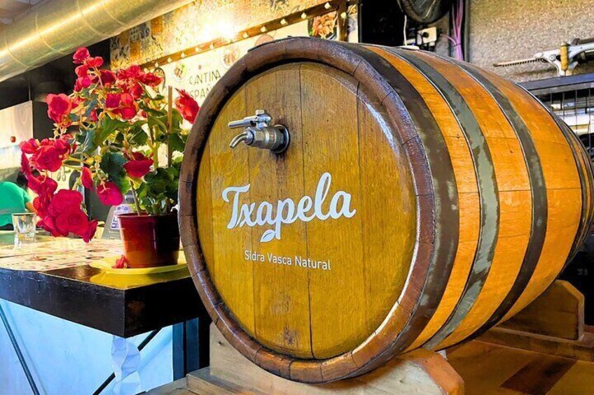 A wooden barrel labeled 'Txapela' with a tap, a potted plant with red flowers, and a counter. 4