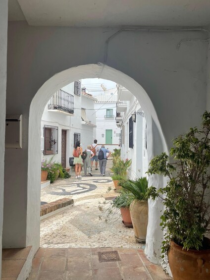 A narrow alleyway with a white archway, potted plants, and a cobblestone path. 1