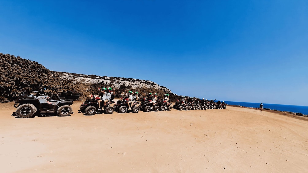 A line of ATVs parked on a sandy beach with a rocky cliff and ocean in the background. 3