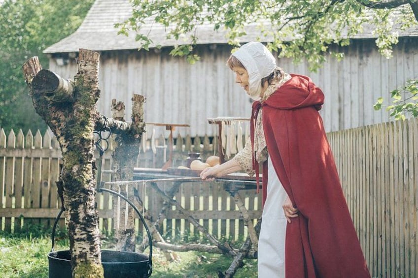 A person in historical attire cooking outdoors with a wood-fired stove and a tree nearby. 3