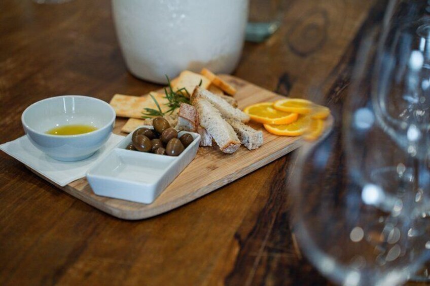 A wooden board with bread, olives, and orange slices, a bowl of olive oil, and a white pitcher on a wooden table. 5