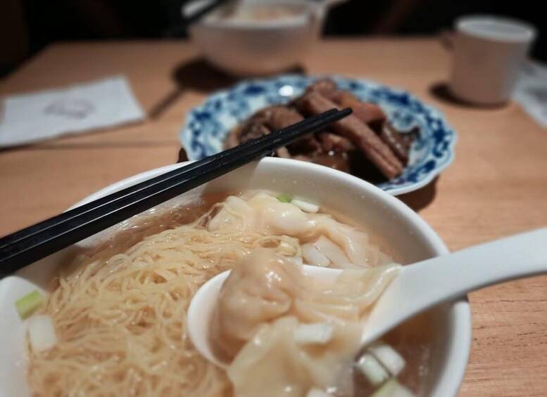 A bowl of noodles with chopsticks and a spoon, a plate with meat, and a cup on a wooden table. 1