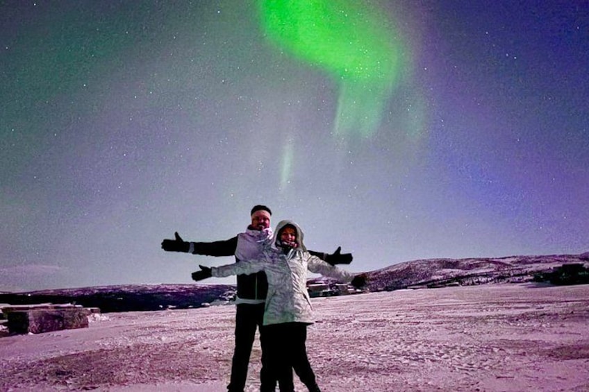 Two people standing on a snowy landscape with a green aurora in the sky. 4