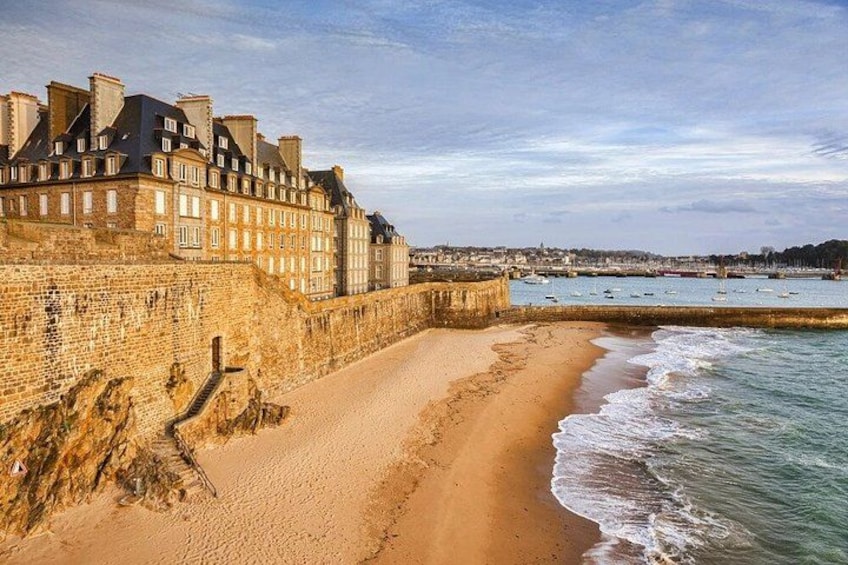 A sandy beach with a stone wall and buildings on the left, and calm sea with boats in the distance. 2