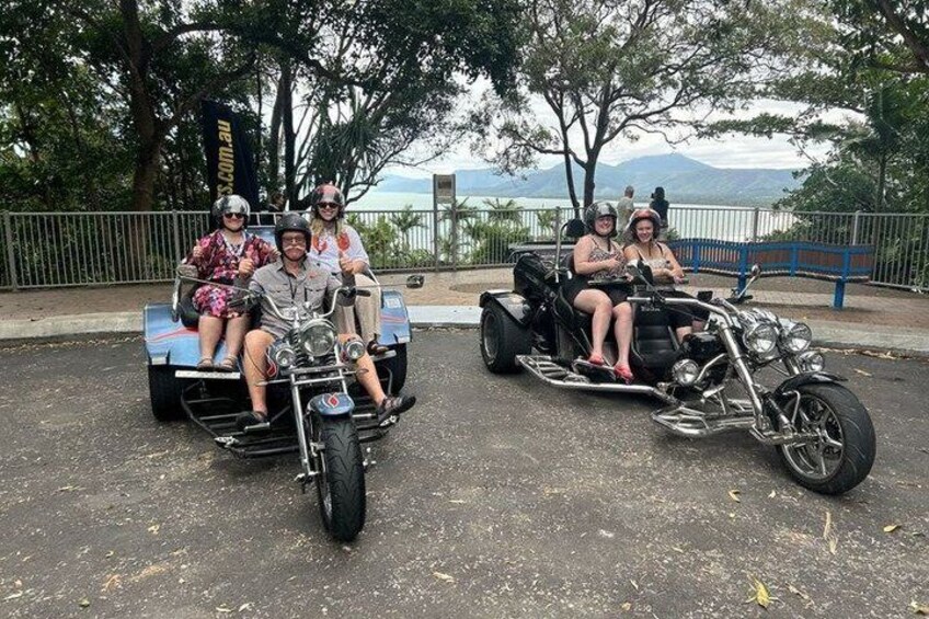 A group of people on a motorcycle and a tricycle, parked on a road with trees and a view of the ocean in the background. 5