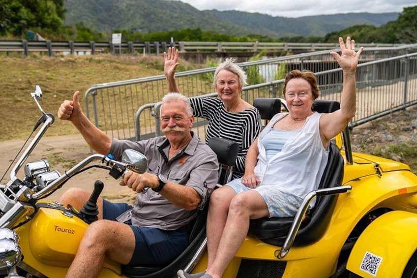 Three people on a yellow vehicle with a metal railing, a body of water, and a bridge in the background. 4