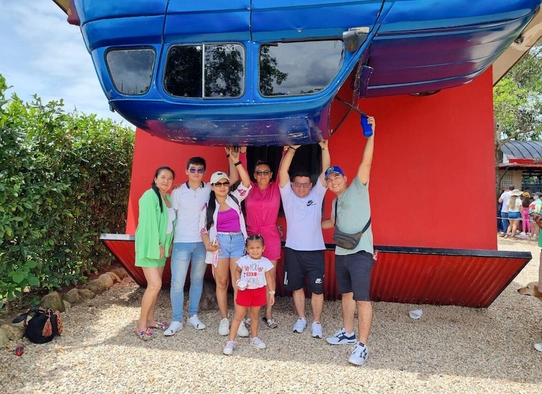 A group of people posing in front of a blue and red structure, possibly a ride or exhibit, with a gravel ground and greenery in the background. 3