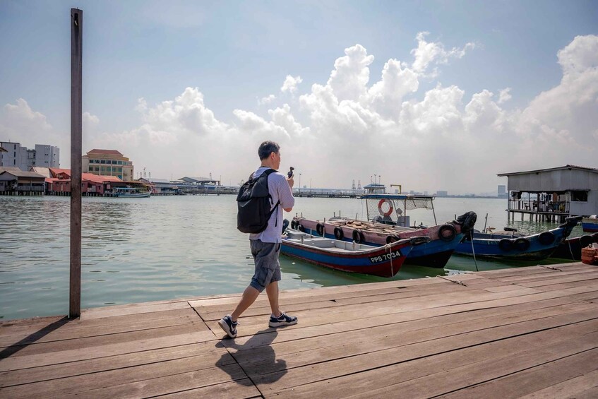 A person walking on a wooden dock towards a body of water with boats and industrial buildings in the background. 3