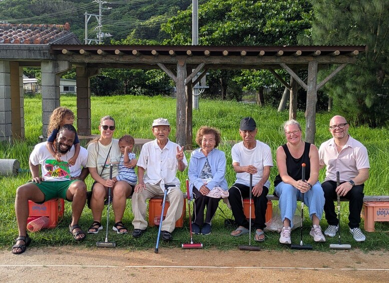 A group of people sitting on a wooden bench under a pergola in a grassy area with trees in the background. 1