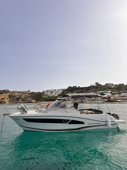 A white yacht named 'Cap Camarat' docked at a marina with clear turquoise water, surrounded by a coastal landscape with buildings and hills. 1