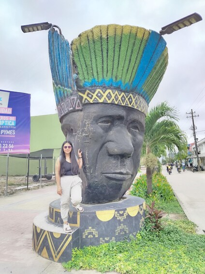 A large sculpture of a face with a headdress, a person standing in front of it, and a street with a banner in the background. 3