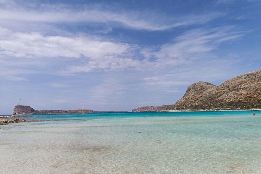 A beach with clear turquoise water, rocky hills, and a distant lighthouse. 1
