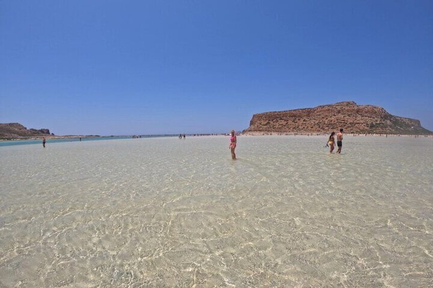 A beach with clear shallow water, a rocky hill in the background, and a few people walking. 4
