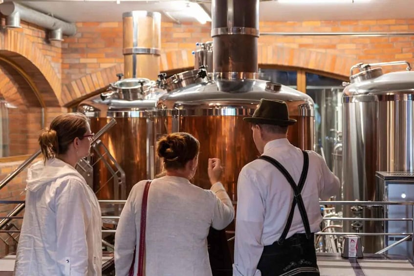 Three people observing industrial brewing equipment in a brick-walled facility. 3
