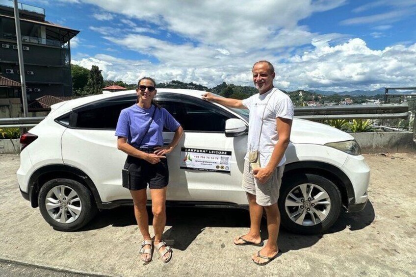 Two people standing next to a white car with a sign on the back. 2